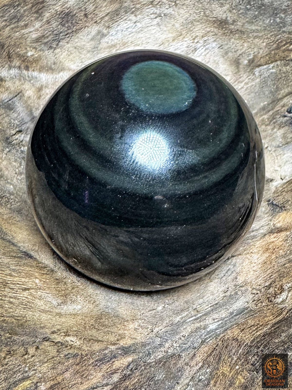 Obsidienne œil céleste inclinée sur bois, œil bleu-vert en partie haute et couronnes vert sombre sur une surface noire polie.
