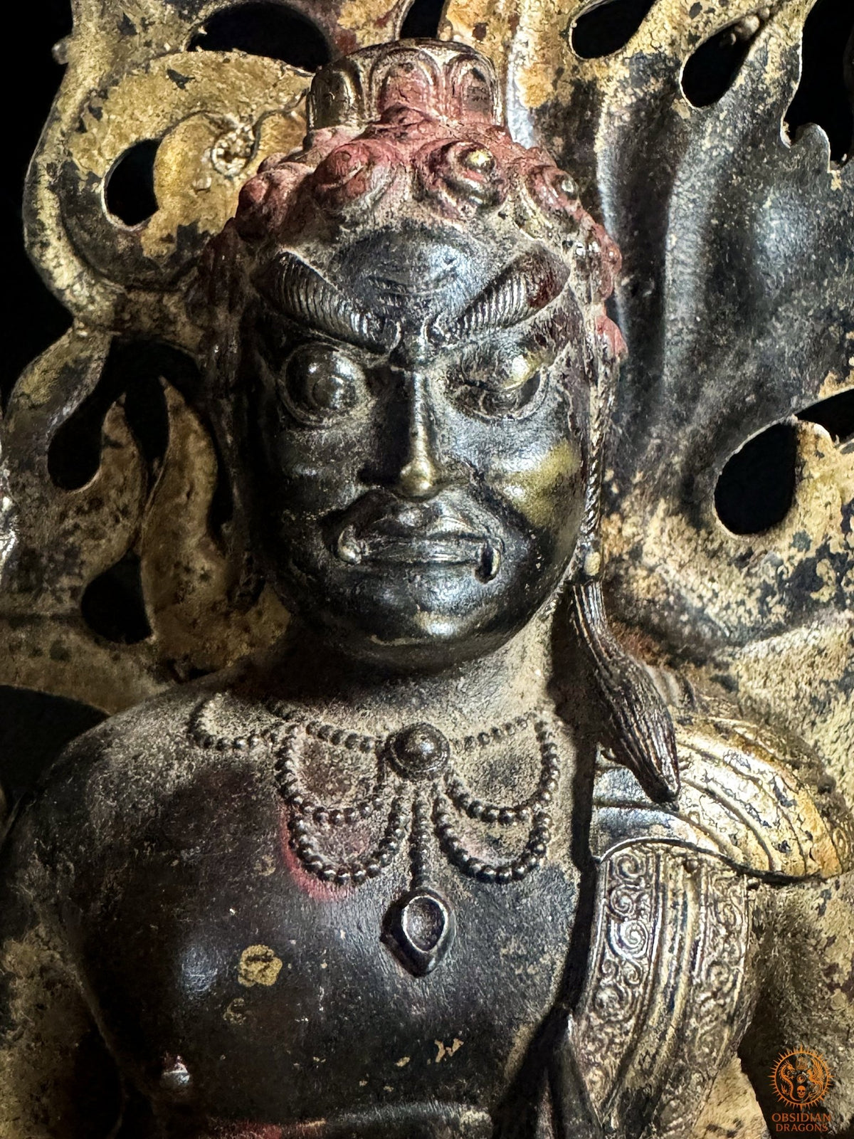 Gros plan sur le visage courroucé d’Acala en bronze, avec yeux globuleux, bouche crispée et patine sombre aux rehauts rouges.