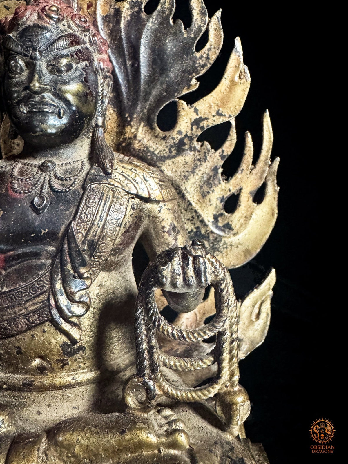 Détail du lasso rituel pāśa dans la main gauche d’Acala, entouré par les flammes dorées de cette sculpture bouddhiste en bronze.