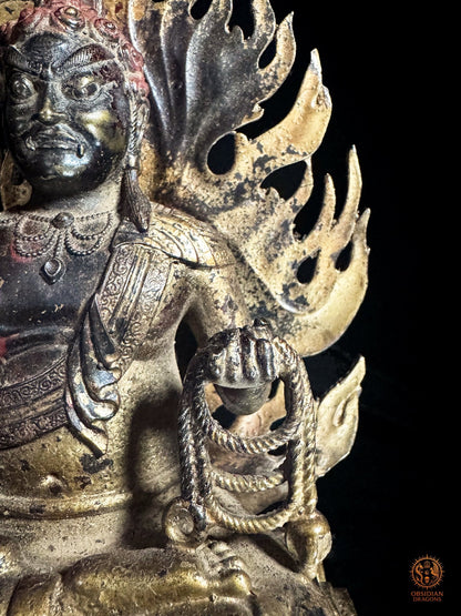 Détail du lasso rituel pāśa dans la main gauche d’Acala, entouré par les flammes dorées de cette sculpture bouddhiste en bronze.