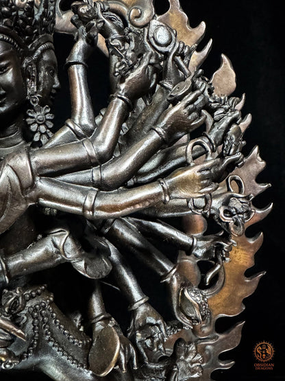 Détail des multiples bras et attributs rituels de Kalachakra sculptés en bronze violet devant la mandorle de flammes ajourée
